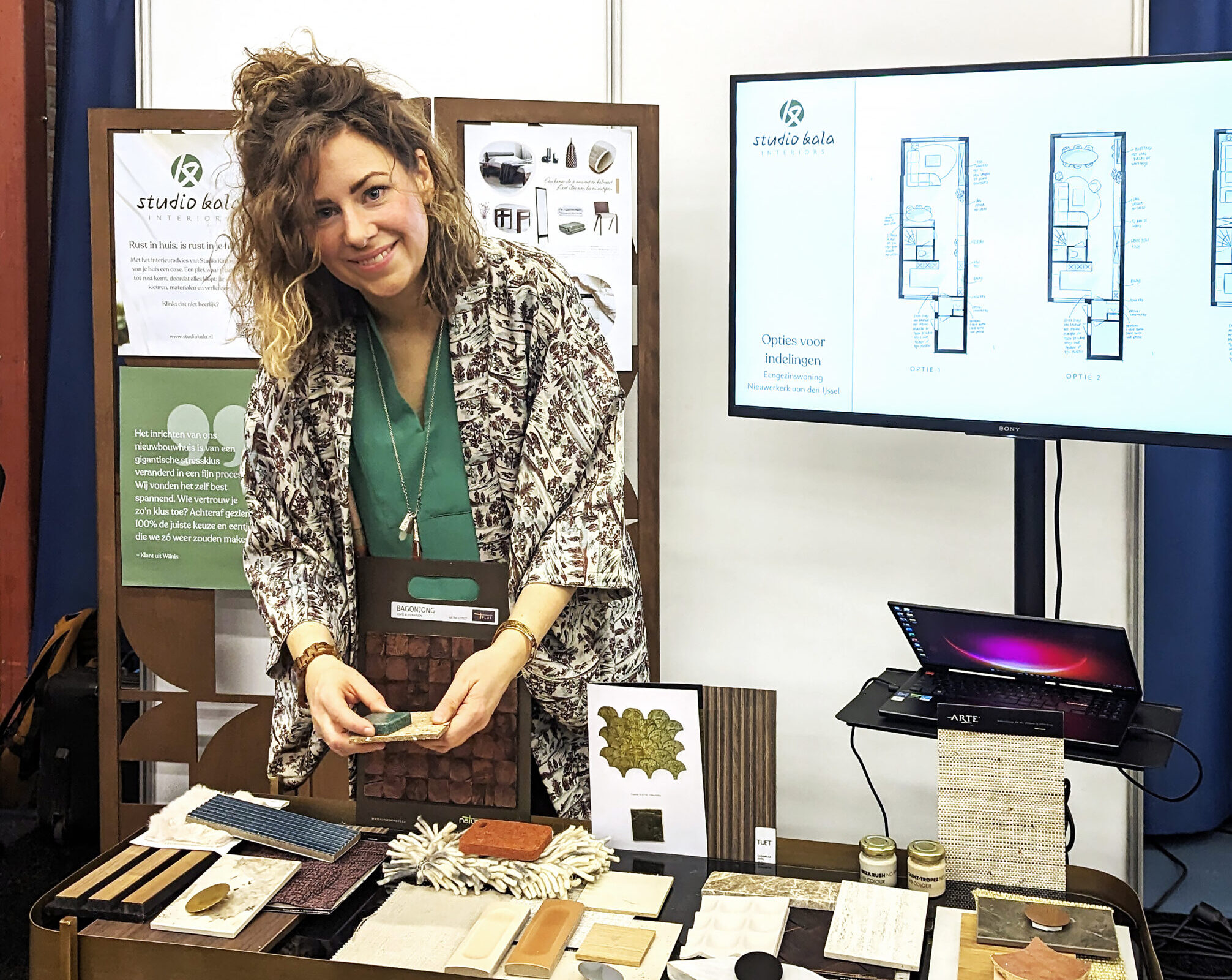 Michèlle van Studio Kala staat bij een tafel vol met materiaalstalen op een beurs. Achter haar een tv scherm waarop schetsen van indelingen van een kamer worden getoond.