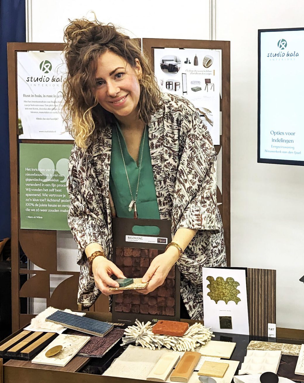 Michèlle van Studio Kala staat bij een tafel vol met materiaalstalen op een beurs. Achter haar een tv scherm waarop schetsen van indelingen van een kamer worden getoond.