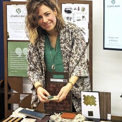 Michèlle van Studio Kala staat bij een tafel vol met materiaalstalen op een beurs. Achter haar een tv scherm waarop schetsen van indelingen van een kamer worden getoond.